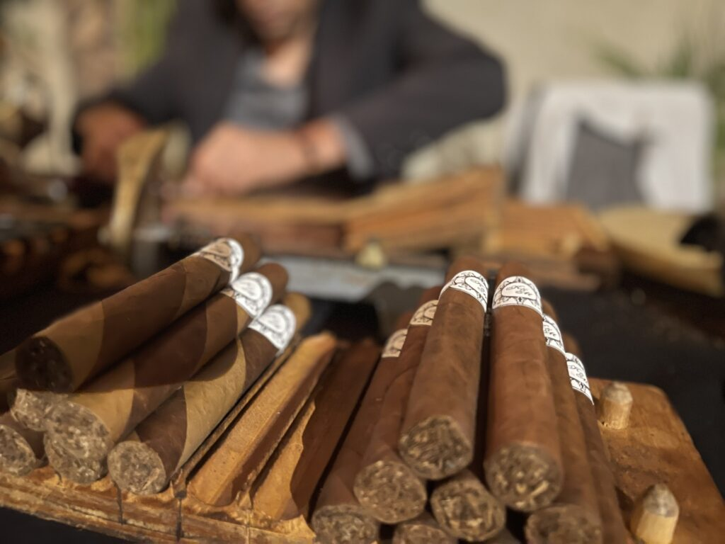 Freshly rolled cigars with bands stacked in the foreground, while a torcedor works at a cigar rolling table in the blurred background.