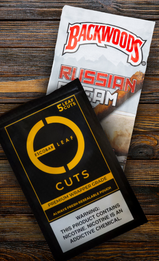 A pack of Escobar Leaf Cuts Premium Wrapper Grade cigar wraps placed over a Backwoods Russian Cream cigar pack on a wooden surface. The Escobar pouch is black and gold with a resealable design and a nicotine warning label, while the Backwoods packaging features its classic logo and cream-toned artwork.