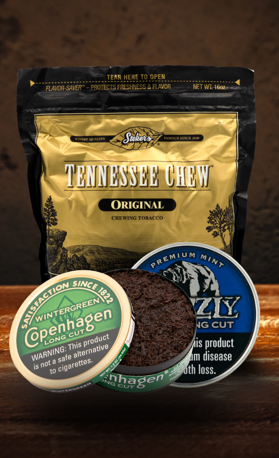 A display of chewing tobacco and dip products on a wooden surface. The background features a gold and black bag of Stoker’s Tennessee Chew Original Chewing Tobacco. In front are two round tins: an open tin of Copenhagen Wintergreen Long Cut showing dark tobacco inside, and a Grizzly Premium Mint Long Cut tin partially visible beside it. The lighting emphasizes the texture of the tobacco and metallic packaging against a dark rustic backdrop.