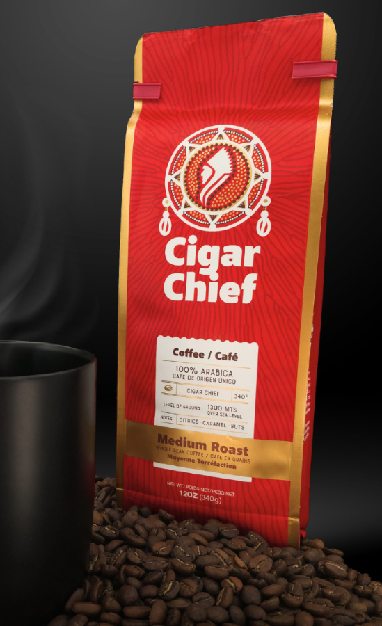 Cigar Chief Coffee — a vibrant red and gold 12oz bag of 100% Arabica medium roast coffee stands upright on a bed of roasted coffee beans beside a matte black mug, with soft smoke rising against a dark background. The packaging features the Cigar Chief logo and gold label detailing caramel and nut flavour notes.